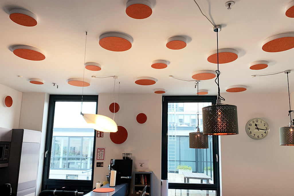 About 20 colored sound absorbers in the form of a large and a small circle (soni ELEMENT CIRCLE) mounted on a ceiling and a wall in the color orange in a white kitchen/breakfast room of a company.