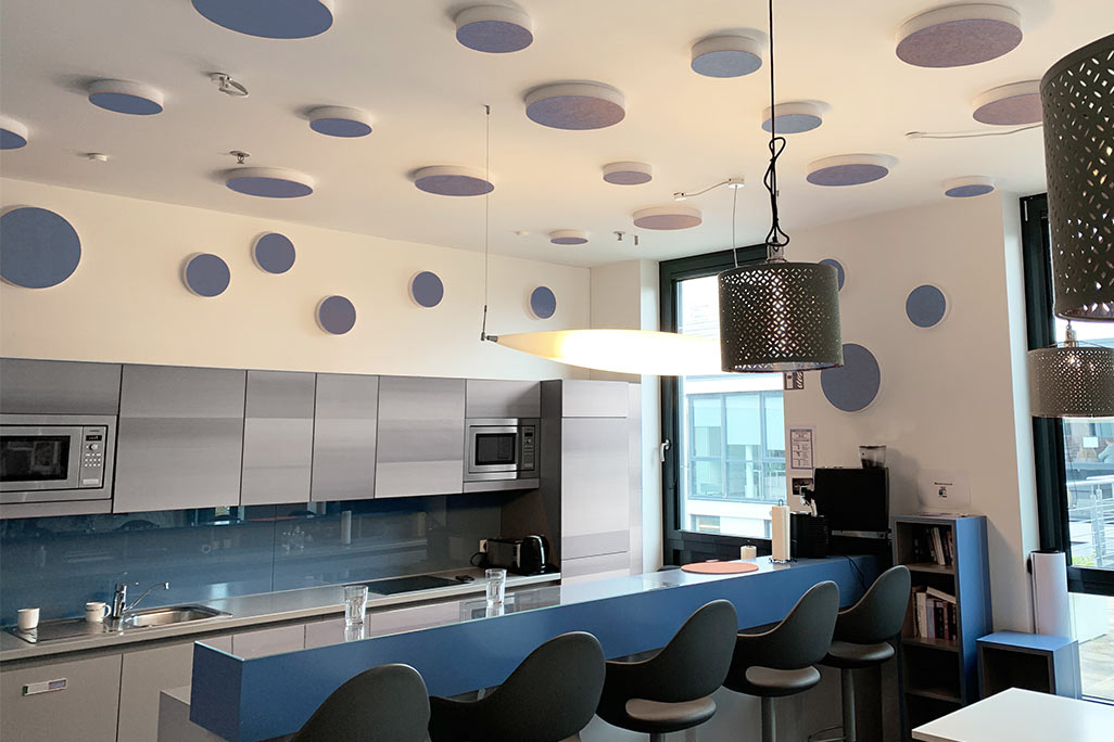 soni ELEMENT CIRCLE About 20 colored sound absorbers in the form of a large and a small circle (soni ELEMENT CIRCLE) mounted on a ceiling and a wall in the color blue in a blue and white kitchen/breakfast room of a company.